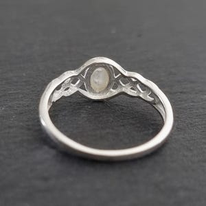 May include: A silver ring with an oval, iridescent gemstone set in a decorative filigree design. The ring band is smooth and polished, and the gemstone appears to be a moonstone. The ring is shown against a dark background.