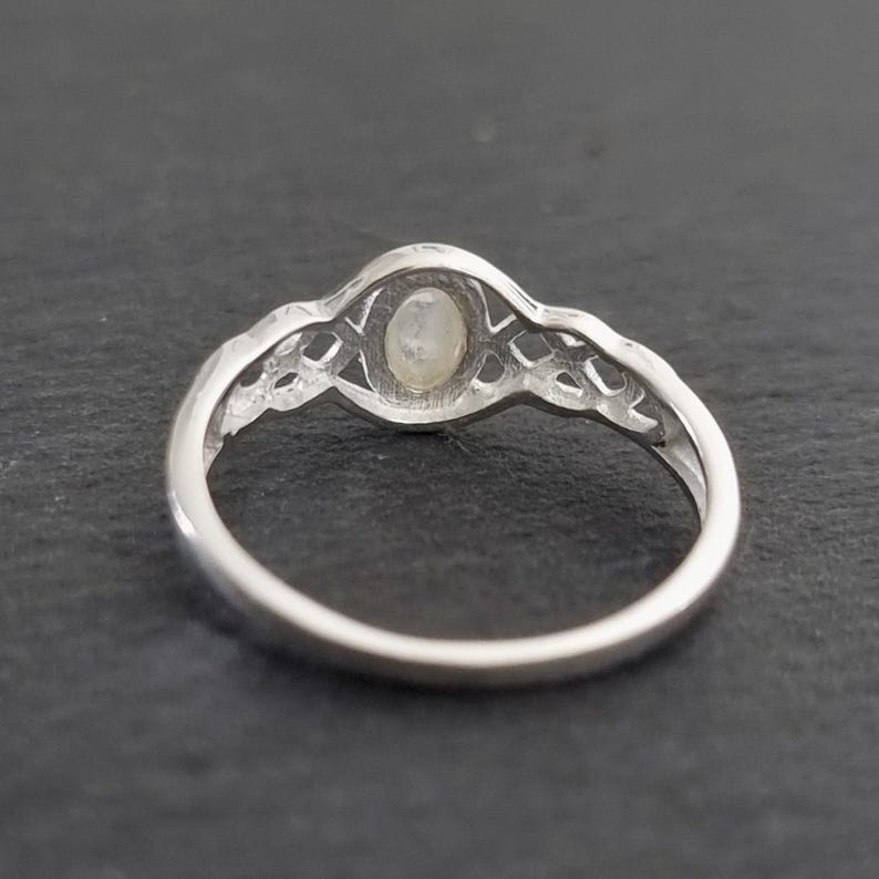 May include: A silver ring with an oval, iridescent gemstone set in a decorative filigree design. The ring band is smooth and polished, and the gemstone appears to be a moonstone. The ring is shown against a dark background.