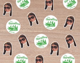 May include: A collection of stickers and die-cut images on a wooden surface. The stickers are circular with a green cabin and text that reads "Camp Caroline". The die-cut images are of a person wearing sunglasses.