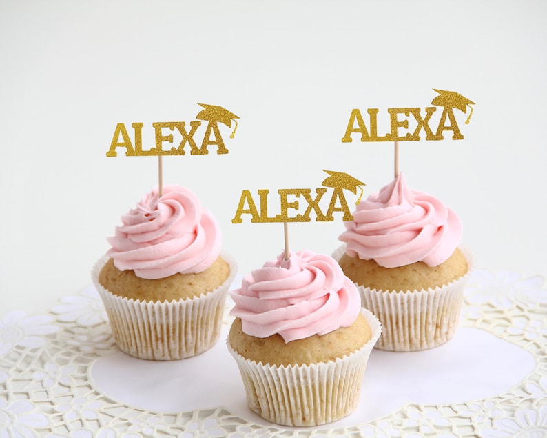 May include: Three cupcakes with pink frosting, each topped with a gold glitter pick that reads "ALEXA" and a graduation cap. The cupcakes sit on a white, lace-edged doily.