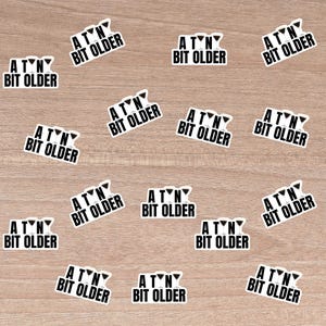 以下が含まれることがあります： 木製の表面に、3つのマティーニグラスのグラフィックと「A Tiny Bit Older」というテキストが書かれた、白黒のステッカーが14枚並んでいます。ステッカーはグリッドパターンで配置されています。