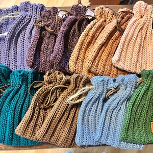 May include: A collection of handmade crochet drawstring bags in various colors, including purple, teal, brown, tan, pink, blue, and green. Each bag features a ribbed texture and a drawstring closure with a leather or cord tie. The bags are arranged on a wooden surface.