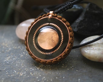 May include: A copper pendant with a black background and a gold trim. The pendant has a copper disc in the center with the letters "LAM" engraved on it. The pendant is on a black cord.