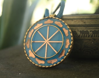 May include: A blue and gold pendant necklace with a circular design featuring a sun symbol with eight rays. The pendant is surrounded by a decorative border.