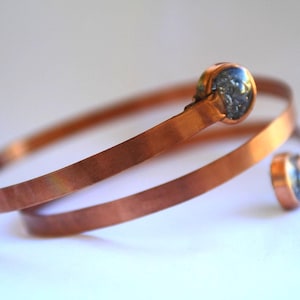 May include: A copper-colored bracelet with a spiral design. The bracelet features two decorative, dark-colored stone accents. The metal has a polished finish, and the bracelet is displayed on a white surface.