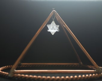 May include: A clear crystal star of David hangs from a copper pyramid on a black background.
