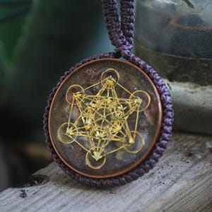 May include: A round pendant with a gold geometric design encased in clear resin. The resin has a purple and brown marbled appearance. The pendant is attached to a purple cord.