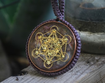 May include: A round pendant with a gold geometric design encased in clear resin. The resin has a purple and brown marbled appearance. The pendant is attached to a purple cord.