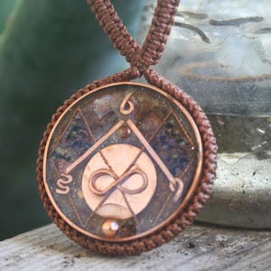 May include: A copper pendant with an infinity symbol and a geometric design, set in a clear resin with iridescent glitter. The pendant is attached to a brown cord.