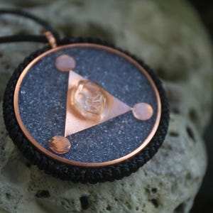 May include: A copper and black pendant with a clear crystal in the center. The pendant is in the shape of a triangle with three circles around it. The pendant is on a black cord.