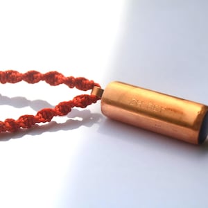 Puede incluir: Un collar con colgante cilíndrico de color cobre con el texto "432 Hz" grabado en el lateral. El colgante está sujeto a un collar de cordón trenzado rojo con dos cuentas doradas.
