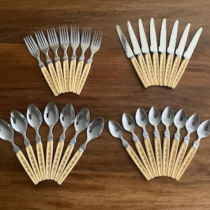 May include: A collection of vintage silverware, including forks, knives, and spoons, arranged on a wooden surface. The silverware features silver blades and tines with cream-colored handles adorned with a gold pattern. The arrangement showcases the cutlery in a fan-like display.