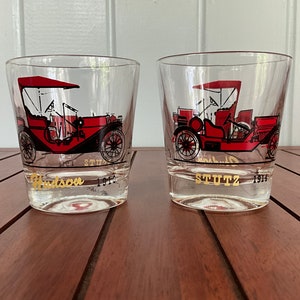 May include: Two vintage glass tumblers with red and black illustrations of antique cars. The left tumbler features a Hudson car from 1910 and the right tumbler features a Stutz car from 1914.