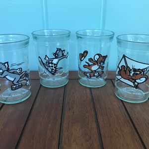 May include: Four clear glass drinking glasses with Tom and Jerry cartoon characters on them. The characters are depicted doing various activities, such as skateboarding, surfing, and flying a kite. The glasses have the Welch's logo on them.