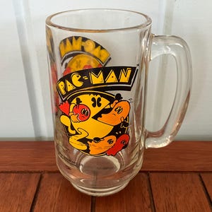 May include: A clear glass mug with a black and yellow Pac-Man graphic. The mug has a handle and says "Pac-Man" in black letters.