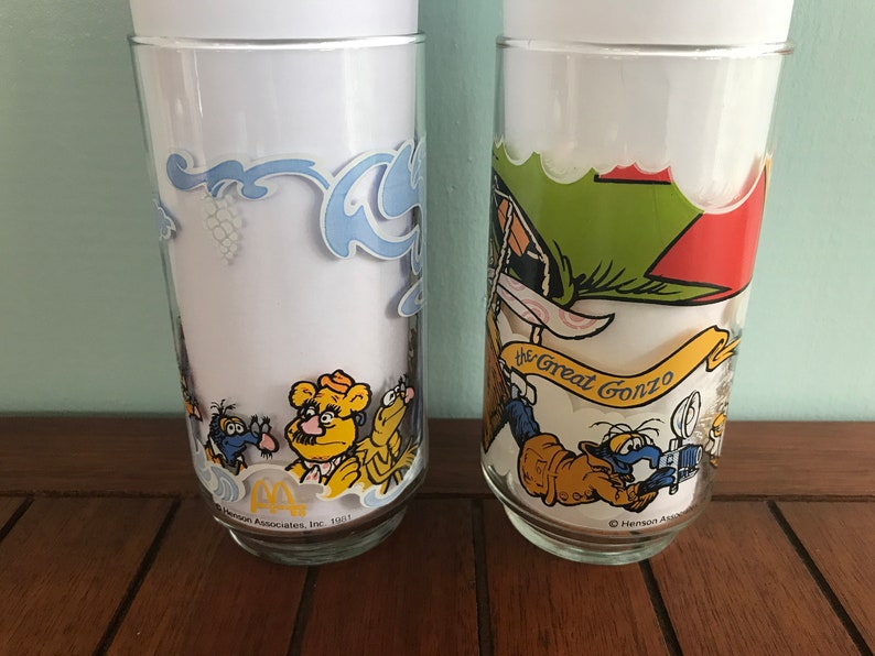 May include: Two vintage glass drinking glasses with colorful cartoon characters. The first glass features a yellow bear with a mustache and a blue bird. The second glass features a blue bird with a red hat and the text "The Great Gonzo".