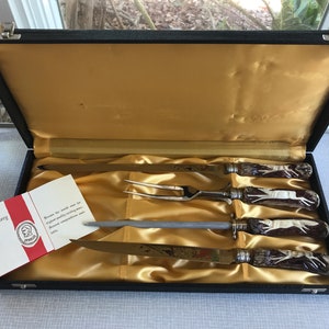 May include: A set of four silver-plated steak knives with stag horn handles in a black velvet-lined box. The knives are arranged in the box with a sharpening steel and a carving fork. The box has a label that reads "Ottella Cutlery".