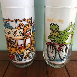 May include: Two vintage glass drinking glasses with colorful cartoon illustrations. One glass features a yellow bus with the text "Happiness Hotel" and various Muppet characters. The other glass features Kermit the Frog riding a bicycle with the text "Kermit the Frog".