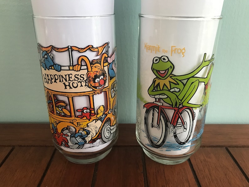 May include: Two vintage glass drinking glasses with colorful cartoon illustrations. One glass features a yellow bus with the text "Happiness Hotel" and various Muppet characters. The other glass features Kermit the Frog riding a bicycle with the text "Kermit the Frog".