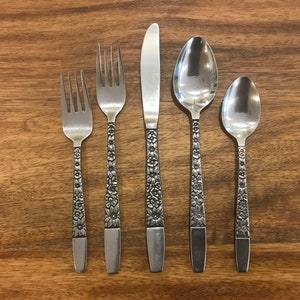 May include: A set of five silver-toned flatware pieces with floral designs. The set includes a knife, two forks, and two spoons.