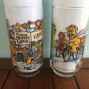 May include: Two vintage glass tumblers with Muppet characters. The first tumbler features Miss Piggy, Kermit the Frog, and other Muppets on a yellow bus with the text "The Great Muppet Caper!" The second tumbler features Fozzie Bear riding a bicycle with other Muppets on bicycles. The text on the bottom of the second tumbler reads "&copy; Henson Associates, Inc. 1981."