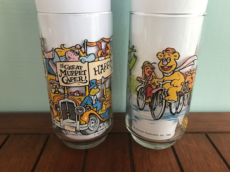 May include: Two vintage glass tumblers with Muppet characters. The first tumbler features Miss Piggy, Kermit the Frog, and other Muppets on a yellow bus with the text "The Great Muppet Caper!" The second tumbler features Fozzie Bear riding a bicycle with other Muppets on bicycles. The text on the bottom of the second tumbler reads "&copy; Henson Associates, Inc. 1981."