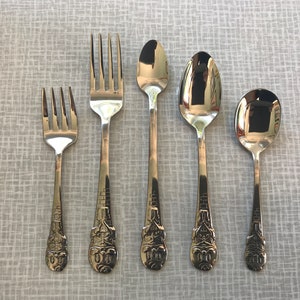 May include: A set of five silver-colored utensils, including two forks and three spoons, arranged on a light gray surface. The handles feature a detailed, ornate design. The spoons vary in size, and the forks have four and three tines.