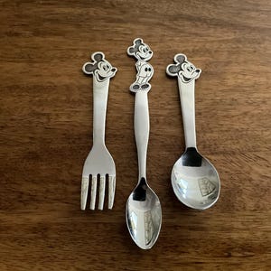 May include: Three silver-colored children's utensils with Mickey Mouse head designs. The set includes a fork, a spoon, and a knife. The utensils are placed on a dark brown wooden surface.
