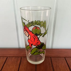 May include: A clear glass featuring a comic book illustration of a red-shirted character with green gloves and pants. The glass has the word "AQUAMAN" in green and white lettering, with a red and green graphic.