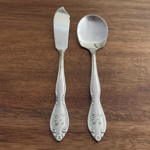 May include: A silver-toned metal butter knife and a matching spoon, both featuring an ornate handle design with an engraved letter K at the base. The utensils have a polished, reflective finish and a decorative floral motif at the very bottom tip.