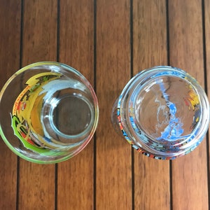 May include: Two clear glass tumblers with colorful designs. The tumbler on the left has a yellow, green, and blue design. The tumbler on the right has a blue, yellow, and red design.