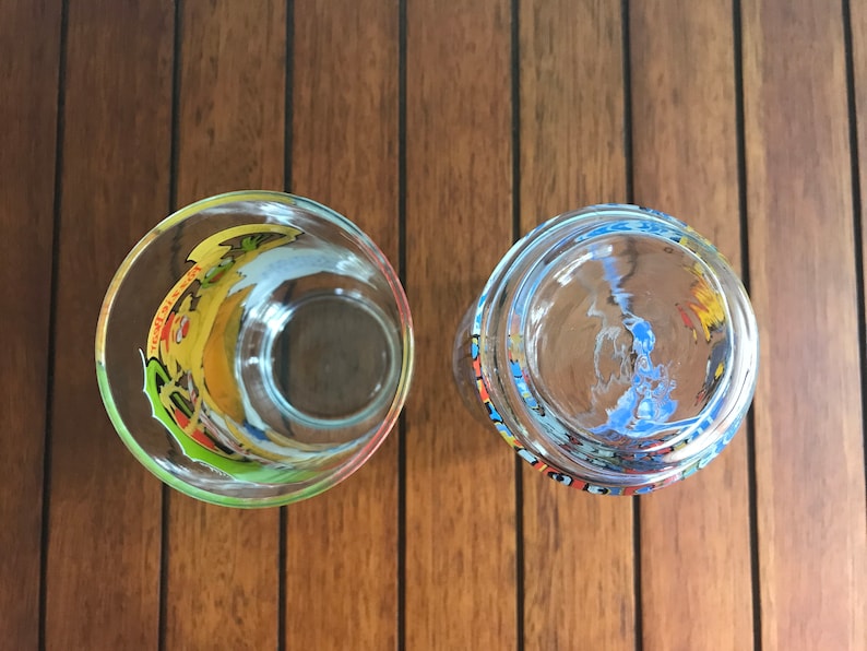 May include: Two clear glass tumblers with colorful designs. The tumbler on the left has a yellow, green, and blue design. The tumbler on the right has a blue, yellow, and red design.