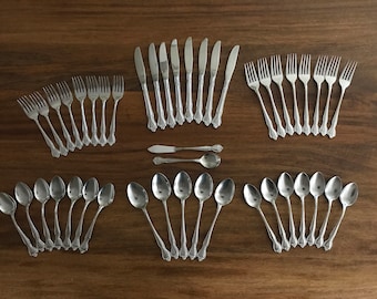 May include: A set of silverware, including forks, knives, and spoons, all made of stainless steel. The silverware is arranged in rows on a wooden surface.