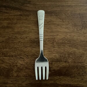 May include: A silver fork with a cartoon chef design on the handle. The handle is engraved with the words "Mmm Good".