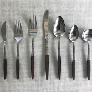 May include: A set of seven silver-toned flatware pieces with brown wooden handles. The set includes a butter knife, two forks, a dinner knife, a soup spoon, a teaspoon, and a dessert spoon.
