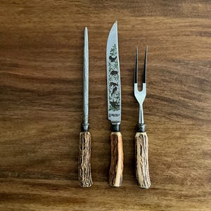 May include: A three-piece carving set with a sharpening steel, knife, and fork. The knife blade features a green and white design with animal silhouettes. Each piece has a handle made of a brown, textured material.