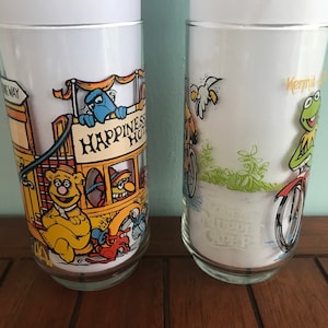 May include: Two vintage glass tumblers featuring characters from Sesame Street. The first tumbler features Big Bird riding a bicycle with the text "Kermit's Bike Shop". The second tumbler features Cookie Monster on a yellow bus with the text "Happiness Hotel".
