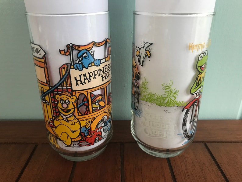 May include: Two vintage glass tumblers featuring characters from Sesame Street. The first tumbler features Big Bird riding a bicycle with the text "Kermit's Bike Shop". The second tumbler features Cookie Monster on a yellow bus with the text "Happiness Hotel".