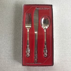 May include: A red box with a set of silverplate cutlery. The box has the text "Campbell Kid Collection" and "International Silverplate". The cutlery includes a fork, knife, and spoon.