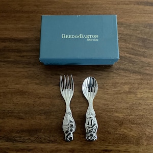May include: A silver fork and spoon set with decorative animal figures on the handles, next to a blue box. The box is labeled "REED & BARTON Since 1824". The set is likely for a child or baby.