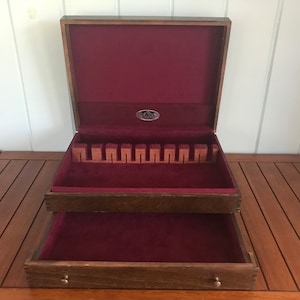 May include: A wooden cutlery box with a burgundy velvet interior. The box has two compartments, one with slots for silverware and the other with a flat bottom.