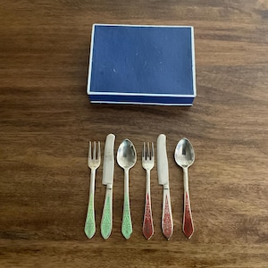 May include: A set of six miniature silver-colored utensils with colored handles, including forks, knives, and spoons, arranged on a wooden surface. A blue box with a white border is in the background. The utensils are likely for a doll or a small tea set.