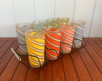 May include: Set of eight striped glass tumblers in a metal holder. The tumblers are in various colors, including blue, green, yellow, orange, and pink. The holder is black metal with a white handle.