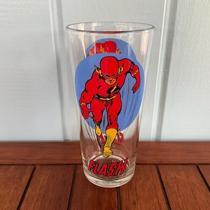 May include: A clear glass featuring the DC Comics superhero, The Flash. The glass has a blue background with a red and yellow Flash graphic. The word "FLASH" is printed in red at the bottom of the glass.