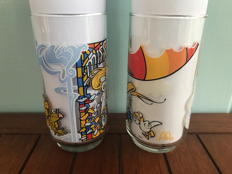 May include: Two vintage McDonald's glasses with cartoon characters. The first glass features a stained glass design with a yellow bird and the text "Mister McFeely". The second glass features a yellow bird flying with a red and yellow umbrella.