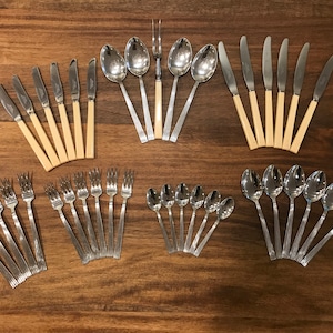 May include: A set of silverware with a mix of forks, spoons, and knives. The silverware is arranged in a fan shape on a wooden surface. The silverware is made of stainless steel and has a polished finish.