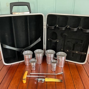 May include: A black aluminum travel bar set with a handle, six silver cups, a corkscrew, and a bottle opener. The set is open and the cups are arranged on a wooden surface.