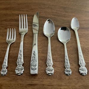 May include: A set of six silver-colored utensils, including forks, knives, and spoons, arranged on a wooden surface. The handles feature a detailed, ornate design. The utensils are likely for dining.