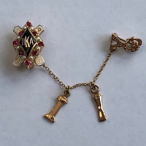 May include: A gold sorority pin with a black diamond and the letters "TIKA" surrounded by red and white stones. A gold chain connects the pin to two gold charms, one shaped like a pillar and the other like a scroll.