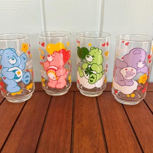 May include: Four clear glass tumblers featuring Care Bears characters in blue, pink, green, and purple. Each glass has a different bear design with hearts and stars. The glasses are on a wooden surface.